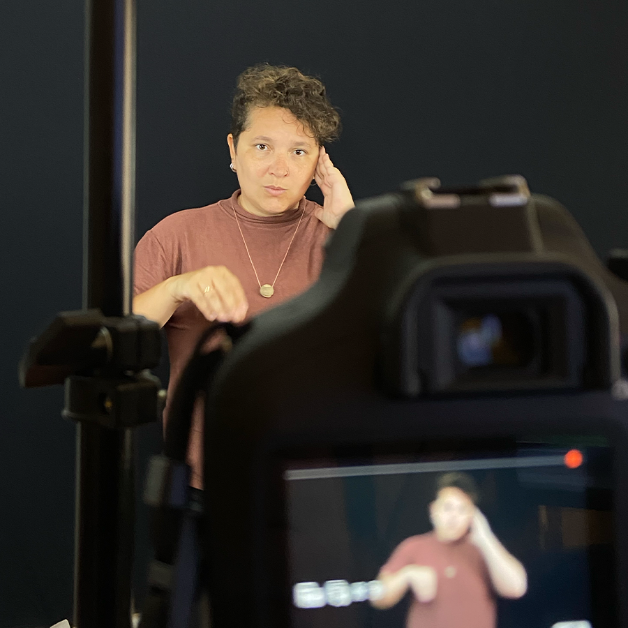 Fotografia de uma pessoa que está de frente para uma câmera fotográfica em gravação. Em um fundo preto, ela sinaliza em Libras.