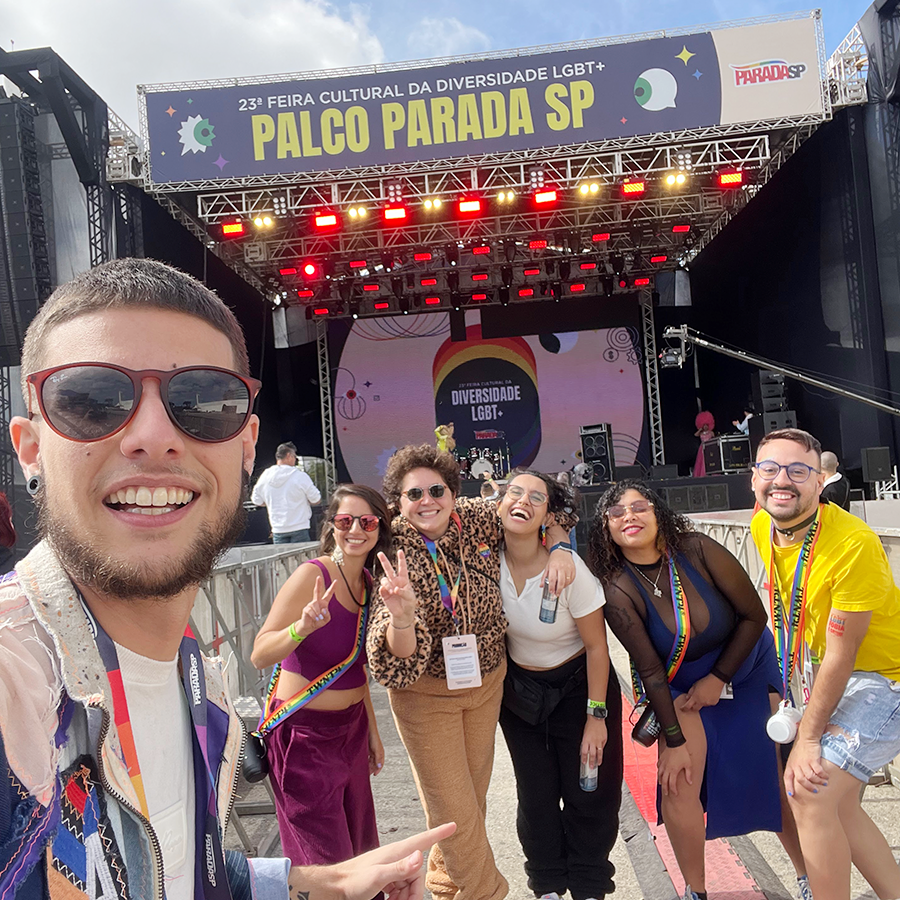 Fotografia de seis pessoas posando para a câmera, sorridentes em um dia de sol. Atrás delas, há um palco com o letreiro PALCO PARADA SP.