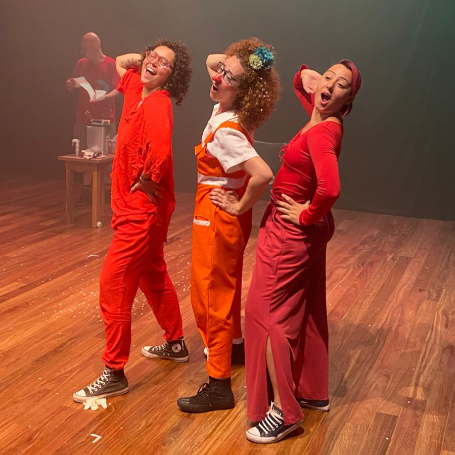 Três pessoas estão posando igualmente de lado e em pé para a câmera. Elas estão apoiando a mão direita sobre a cabeça, boquiabertas e sorridentes. Estão usando roupas vermelhas vibrantes, e a do centro, está usando um nariz de palhaço e com o cabelo enfeitado.