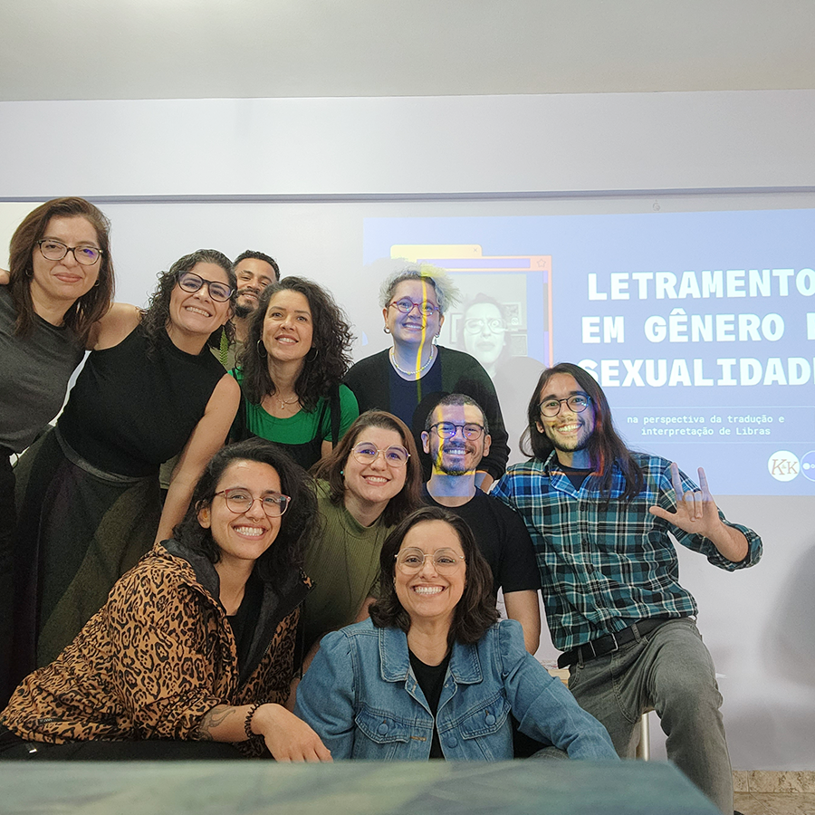 Fotografia de dez pessoas, sorrindo para a câmera. Atrás delas, projeta uma imagem com o texto "Letramento de Gênero e Sexualidade".