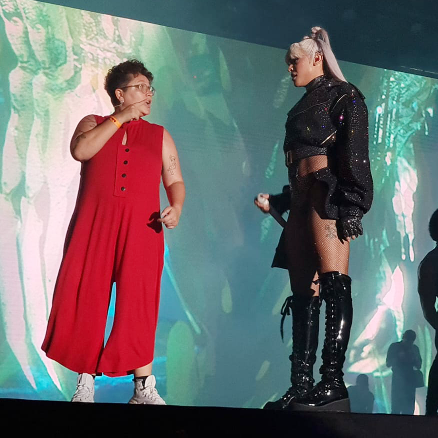 Fotografia de uma mulher vestida de vermelho, sinalizando em Libras. Ela encara Pabllo Vittar, que está retribuindo o olhar.